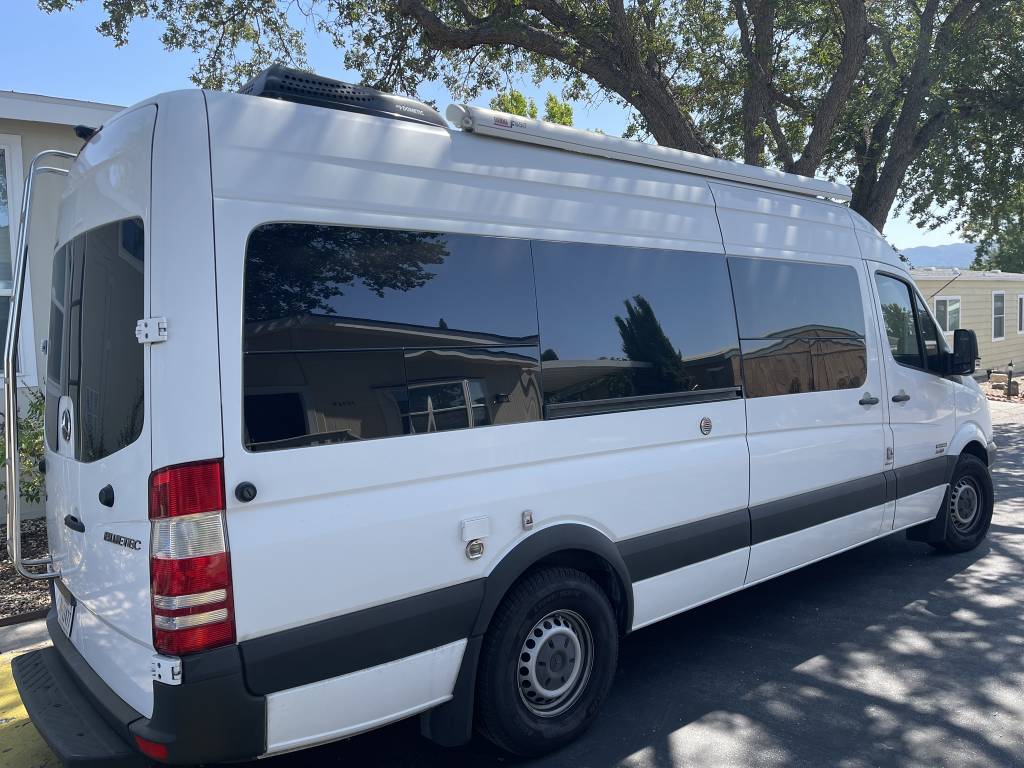 A white Mercedes Sprinter camper van, ideal for rv rental, is parked outdoors. Features tinted windows, rooftop AC, and side awning.