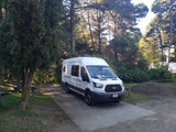 White Ford Transit camper van, an ideal RV rental, parked on a paved site in a sunny forest campground.