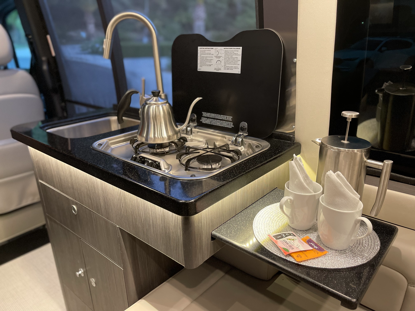 This RV rental kitchenette features dark counters, brushed cabinets, a sink, stovetop with kettle, and a coffee/tea station.