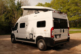 White Winnebago Solis pop-top camper van rental parked on a dirt path amidst trees, perfect for outdoor trips.