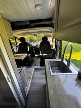 RV rental interior: View from rear towards cockpit, with kitchen sink, wood-look floor, and window coverings visible.