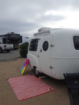 Small white RV rental (travel trailer) set up on a sandy site. Features a colorful boogie board & outdoor rug.