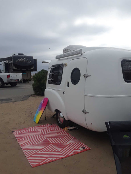 Small white RV rental (travel trailer) set up on a sandy site. Features a colorful boogie board & outdoor rug.