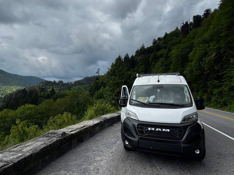 Our white Ram ProMaster RV rental on a mountain road, with cloudy skies and lush forests. Adventure awaits!