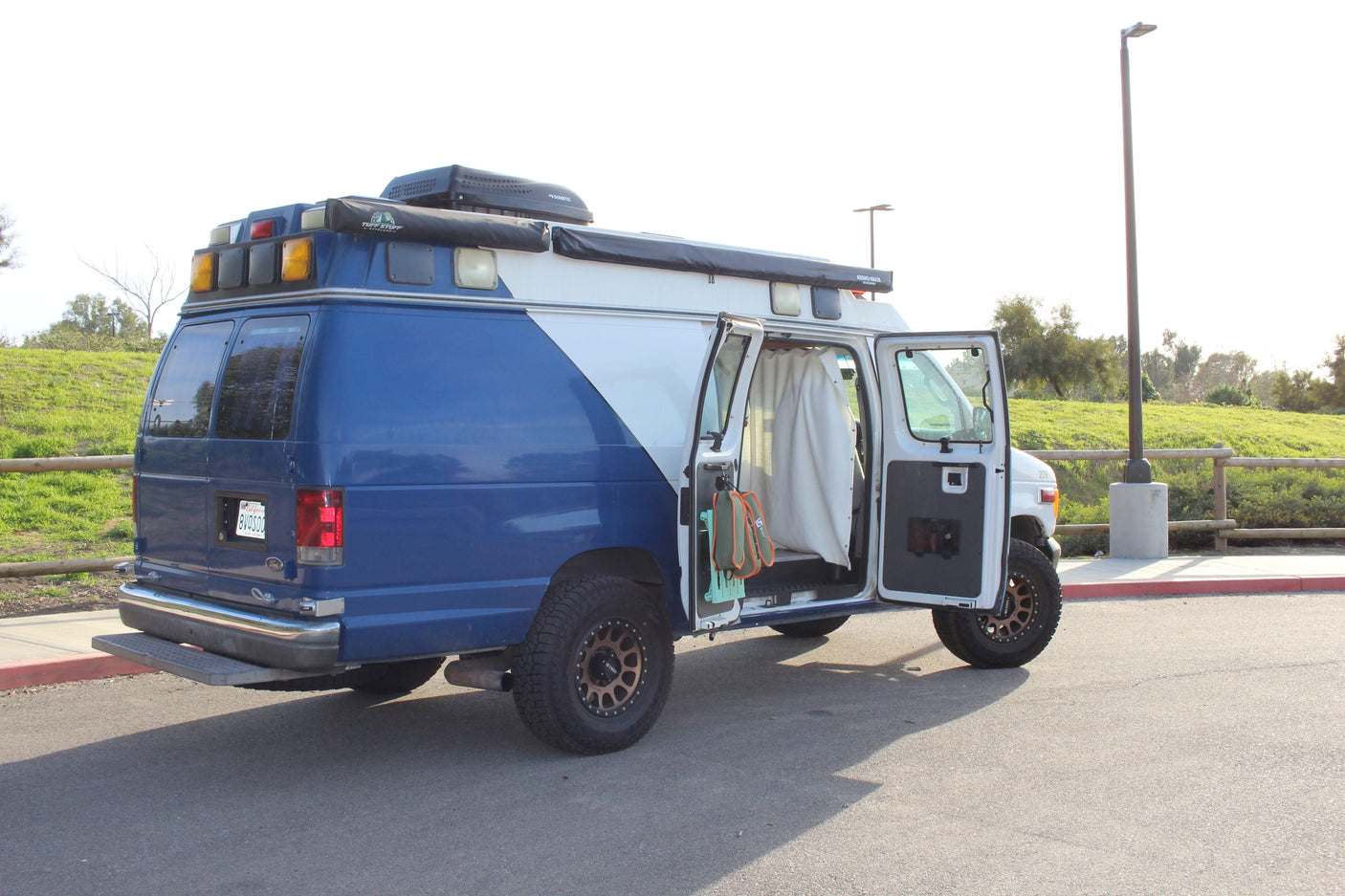 A custom blue/white adventure van, ideal for RV rental, with off-road tires, roof awnings, and an open side revealing its camper interior.