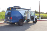 A custom blue/white adventure van, ideal for RV rental, with off-road tires, roof awnings, and an open side revealing its camper interior.