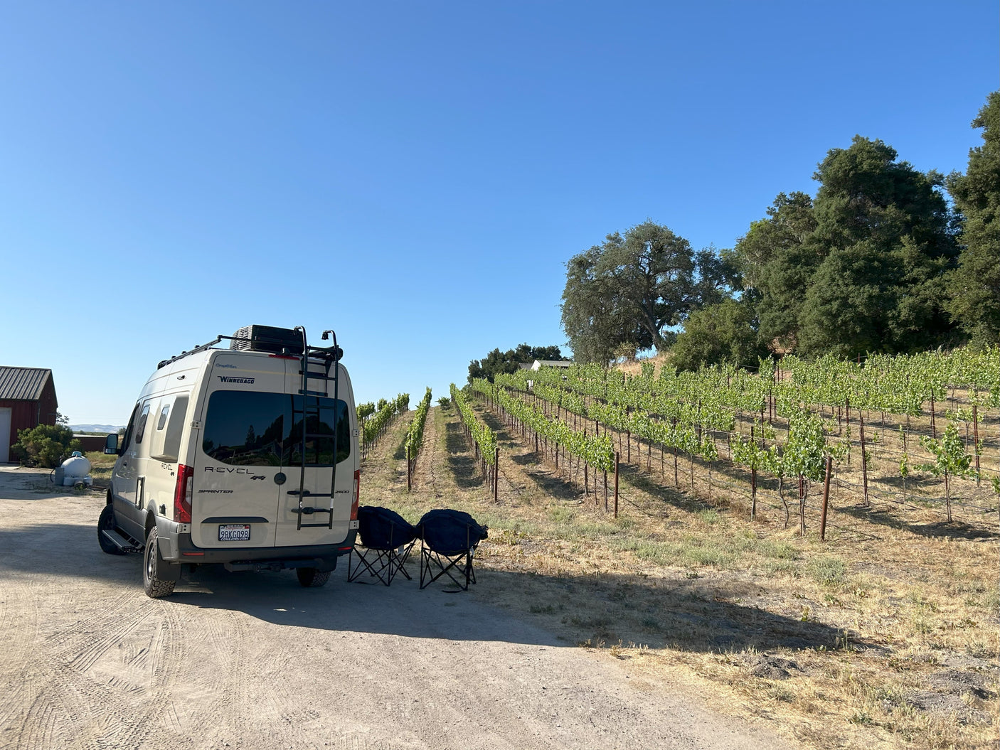 Scenic RV rental adventure: A white Winnebago Revel van is parked by lush vineyard rows, with chairs set for relaxation.