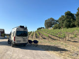 Scenic RV rental adventure: A white Winnebago Revel van is parked by lush vineyard rows, with chairs set for relaxation.