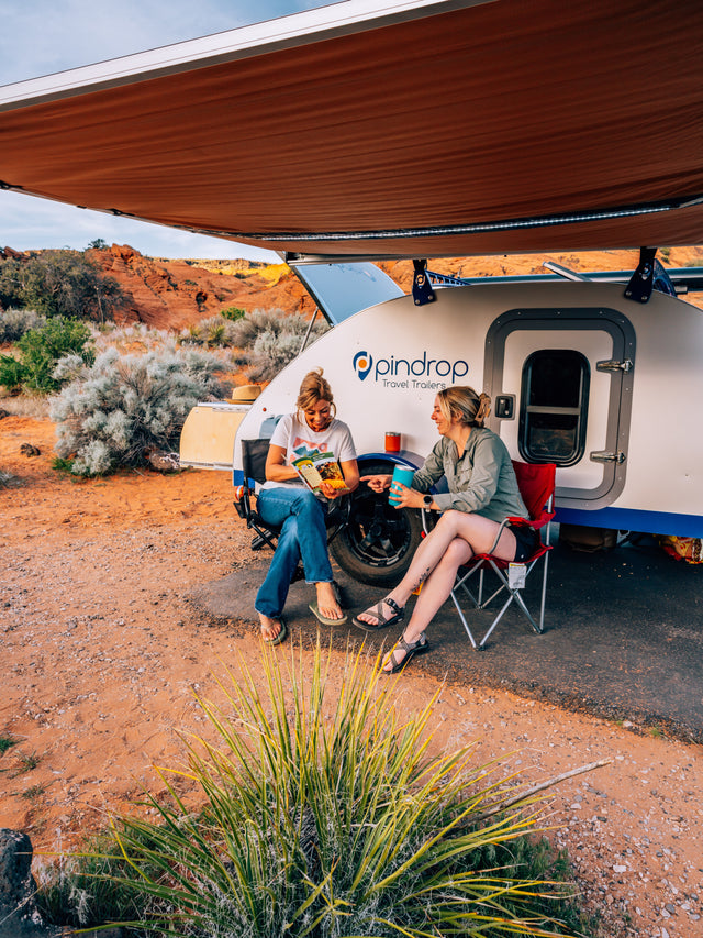 Two women relax under an awning next to a Pindrop rv rental travel trailer in the desert. One reads, the other sips a drink.