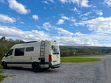 Light-colored Sprinter RV rental parked on gravel. Scenic view of green hills, trees, and blue sky with white clouds.