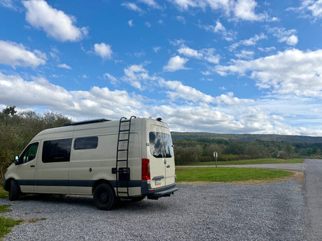 Light-colored Sprinter RV rental parked on gravel. Scenic view of green hills, trees, and blue sky with white clouds.