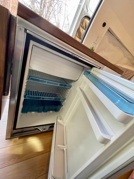 RV rental interior: A compact refrigerator, open to show its freezer compartment, shelves, and door storage inside.