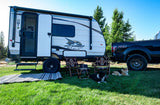 Enjoy a scenic RV rental adventure! This travel trailer is set up with a truck, camping chairs, and two happy dogs at a beautiful outdoor campsite.