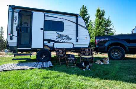 Enjoy a scenic RV rental adventure! This travel trailer is set up with a truck, camping chairs, and two happy dogs at a beautiful outdoor campsite.