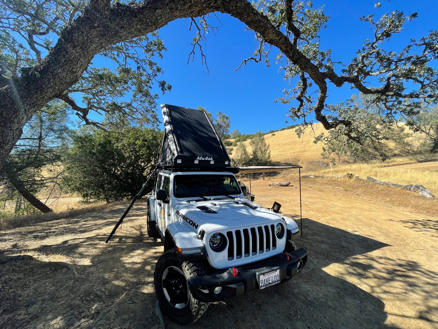 White Jeep Wrangler with rooftop tent & awning under trees, ready for an RV rental camping adventure in dry hills.
