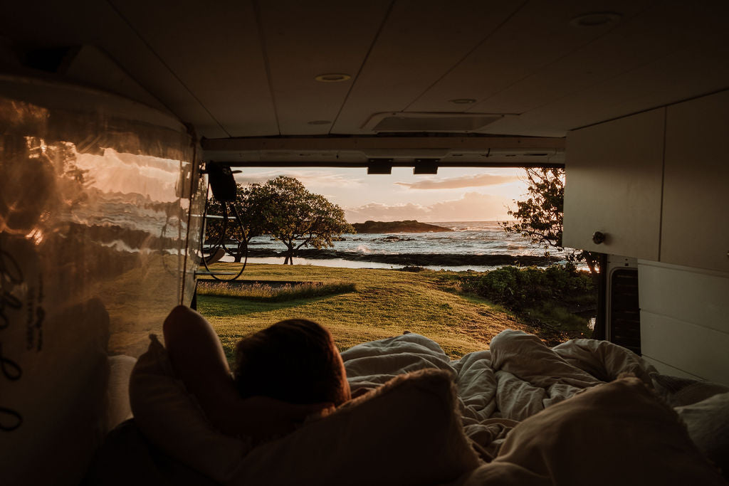 View from inside an RV rental looking out at a sunset over the ocean, with a grassy lawn and trees. A person relaxes in bed.