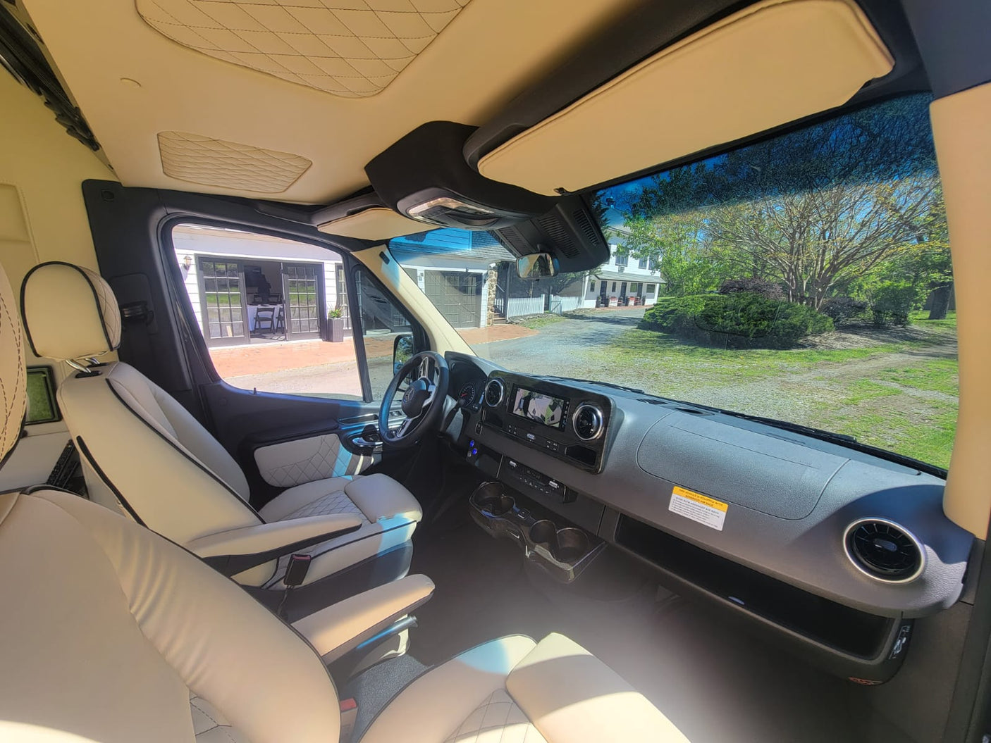 Luxurious RV rental interior: Cream leather seats, detailed dashboard, and front view of a house and trees.