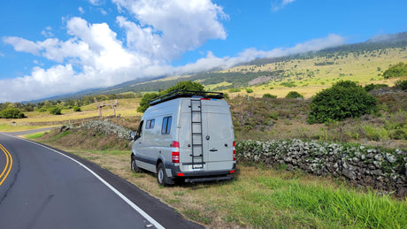 Grey RV rental van on a scenic road trip, parked by a stone wall with misty green mountains.
