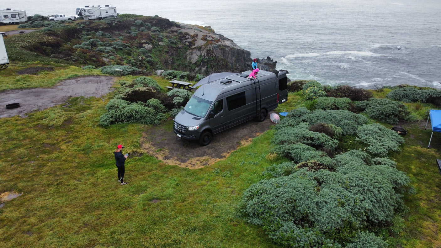Grey RV rental van parked cliffside overlooking the ocean, with children on roof and a person nearby. Coastal camping scene.