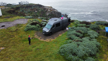 Grey RV rental van parked cliffside overlooking the ocean, with children on roof and a person nearby. Coastal camping scene.