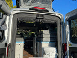 Rear view of an RV rental van interior, featuring a portable toilet, stacked bedding cushions, and storage compartments.