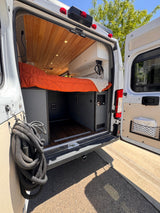 Interior of an RV rental camper van with a cozy bed, wooden ceiling, and storage, seen from the open back doors.