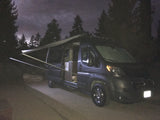 Night shot of an RV rental with its awning extended, parked in a forest. A dog with glowing eyes peeks from the open side door.