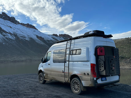 A dirty white rv rental camper van with adventure gear parked lakeside before snowy mountains under a cloudy sky.