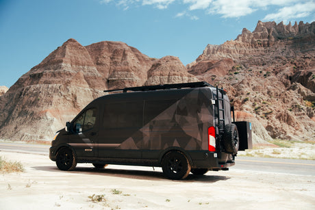 Customized dark RV rental van with a geo pattern and roof rack parked in rugged badlands terrain.