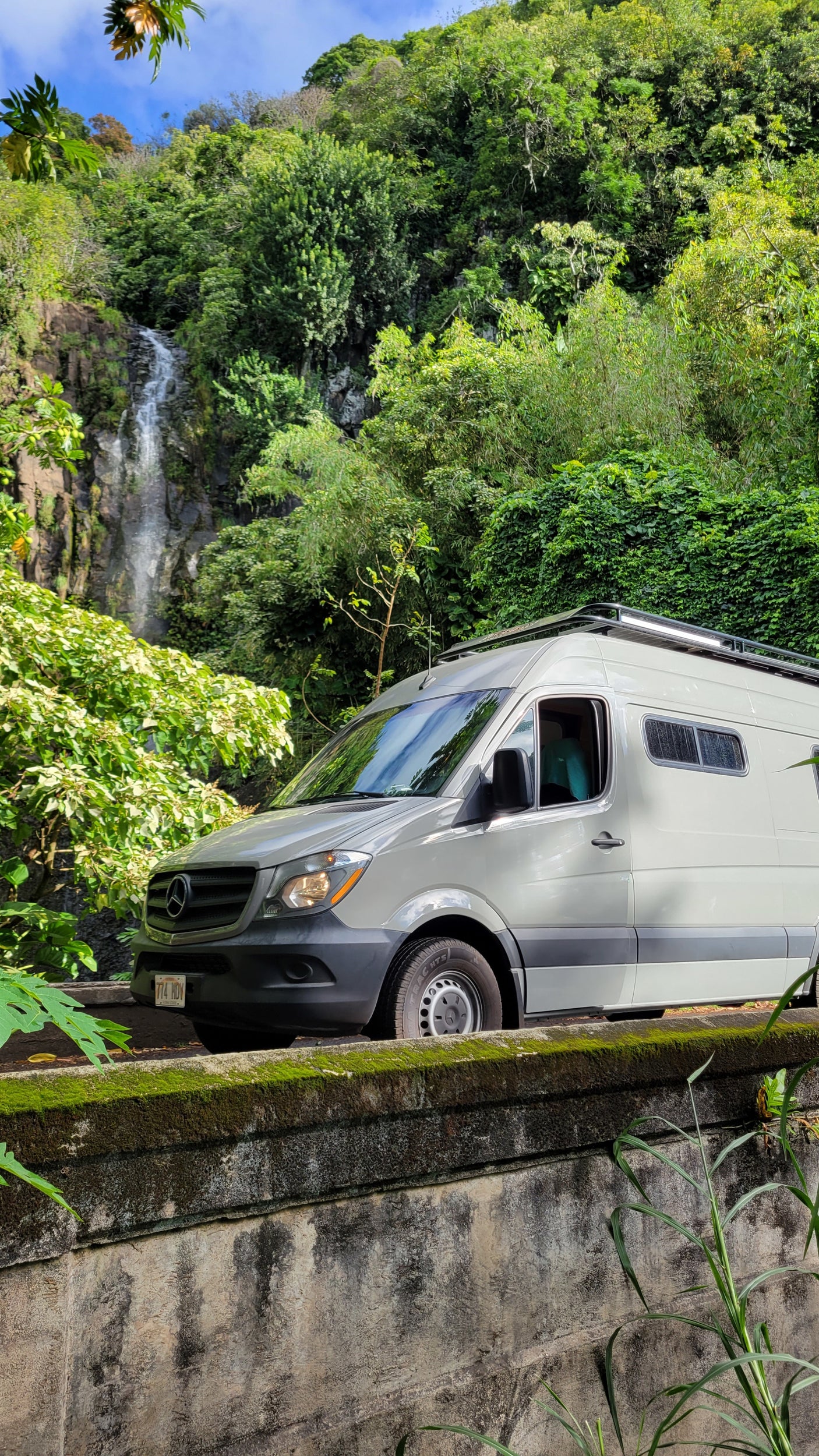Scenic view of a grey Mercedes RV rental van parked on a mossy wall with a waterfall & lush greenery in the background.