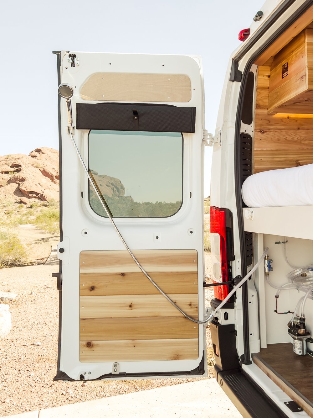 White RV rental van's open rear door shows outdoor shower & interior wood build. Perfect for remote trips.