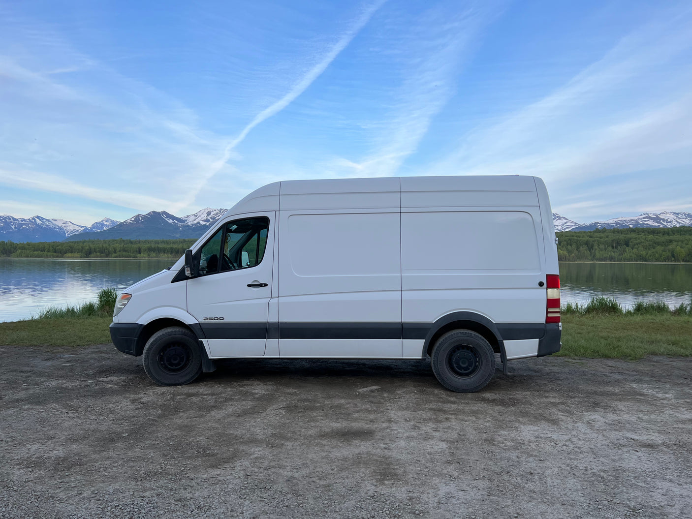 This white Sprinter van, ready for RV rental, is parked by a tranquil lake with snow-capped mountains.