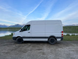 This white Sprinter van, ready for RV rental, is parked by a tranquil lake with snow-capped mountains.