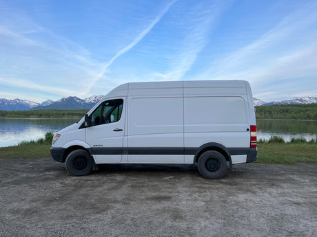 This white Sprinter van, ready for RV rental, is parked by a tranquil lake with snow-capped mountains.