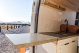 RV rental ready: Spacious camper van interior with a modern kitchenette, showcasing a concrete counter, sink, & mountain views.