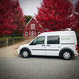 White Maine Campervan Rentals van, an RV rental, parked near a red barn framed by brilliant red autumn trees.