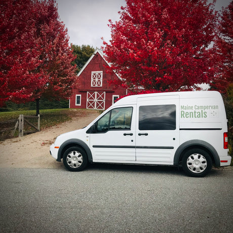 White Maine Campervan Rentals van, an RV rental, parked near a red barn framed by brilliant red autumn trees.