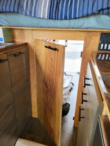 Inside an RV rental: view of a striped bed, teal blanket, wood cabinetry, and a control panel for camper systems.