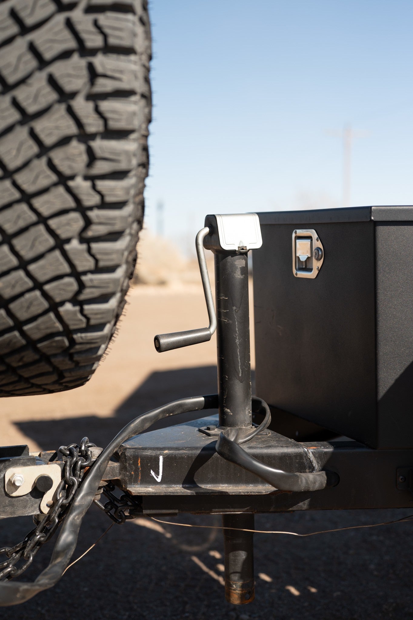 Close-up of an RV rental trailer's hitch, jack, safety chains, and toolbox, with a tire in the foreground.