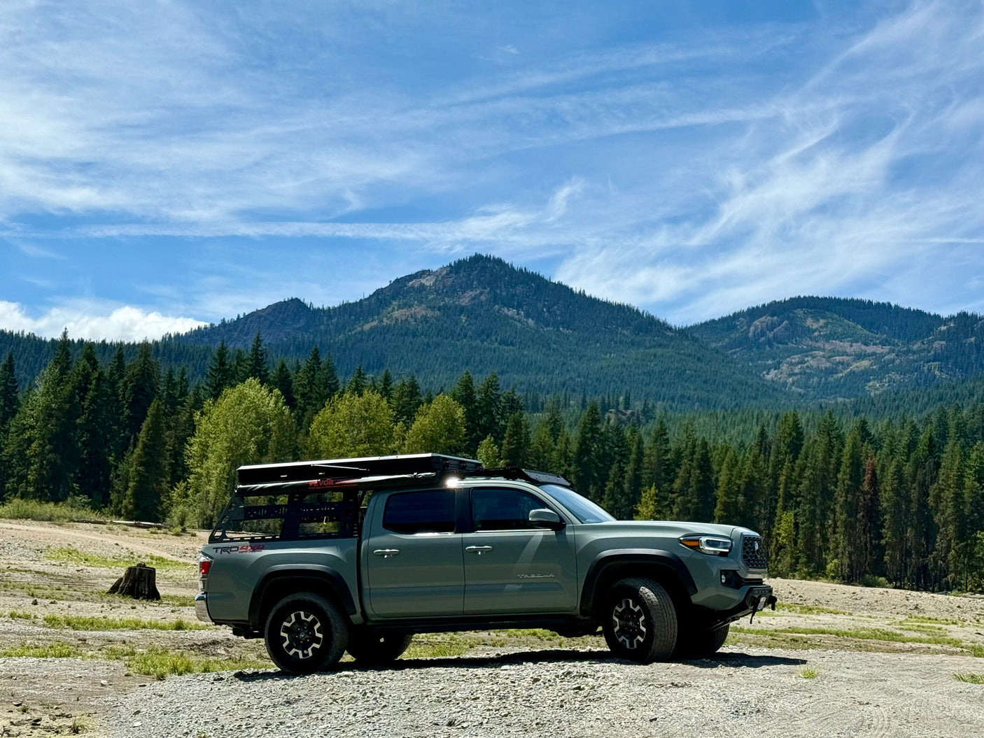 Grey overlanding truck with rooftop tent parked in front of forested mountains. Great for outdoor trips, like an RV rental.