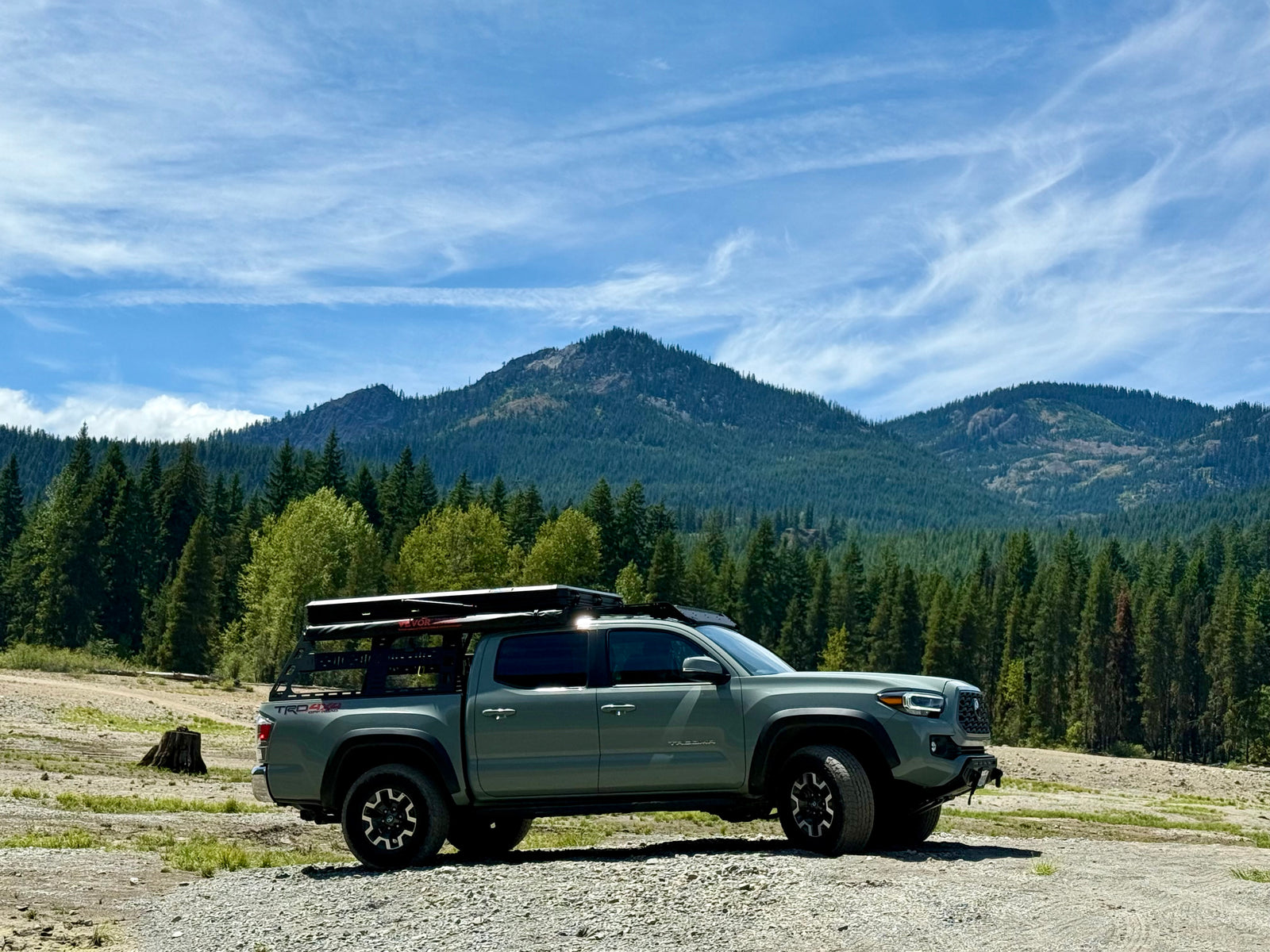 Grey overlanding truck with rooftop tent parked in front of forested mountains. Great for outdoor trips, like an RV rental.