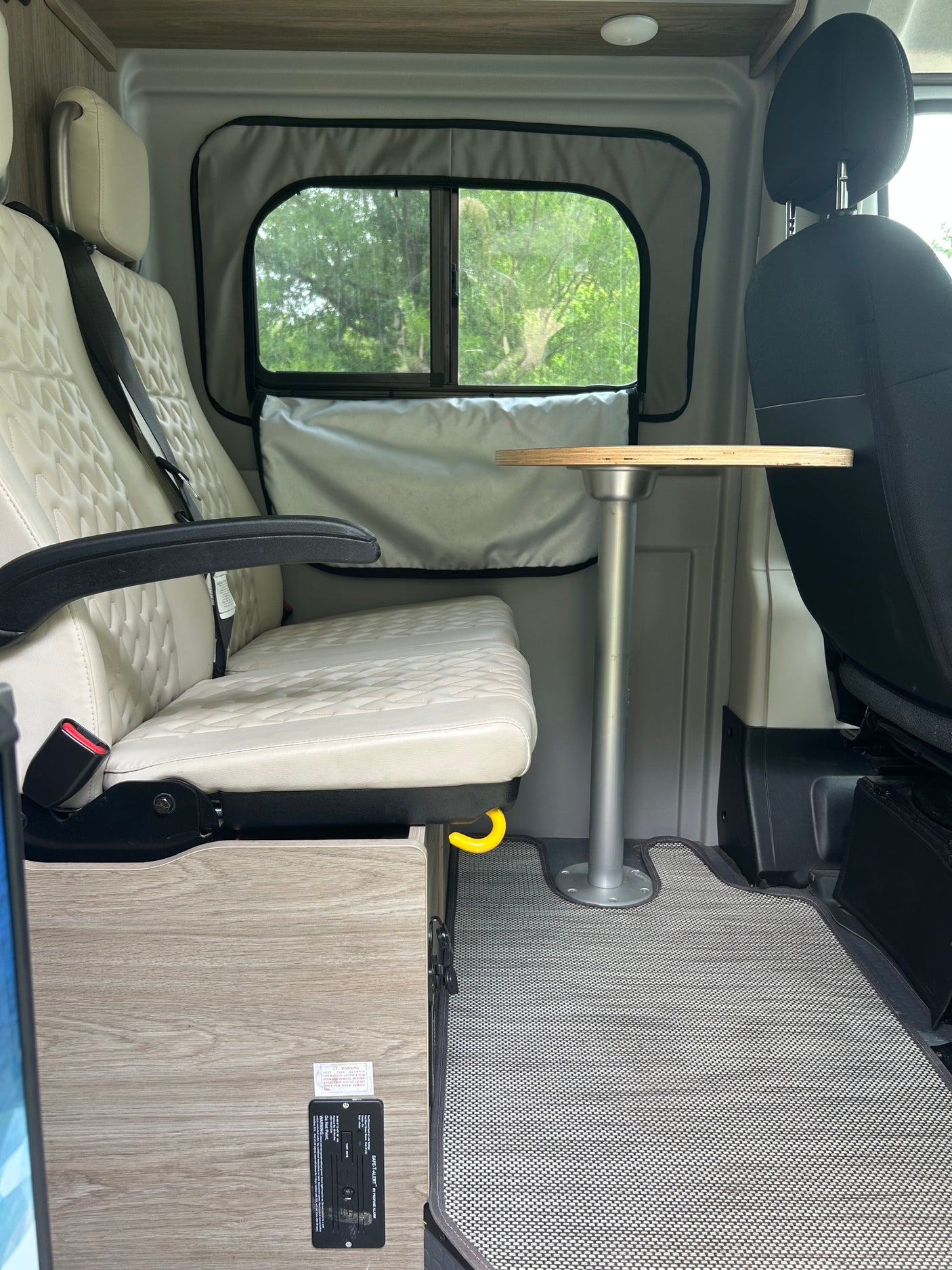 An inviting rv rental interior with quilted beige seats, a wooden table, and a window offering a lush green view.