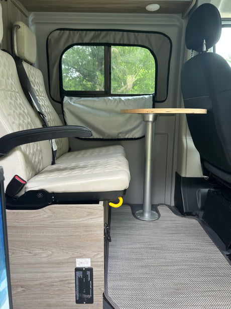 An inviting rv rental interior with quilted beige seats, a wooden table, and a window offering a lush green view.