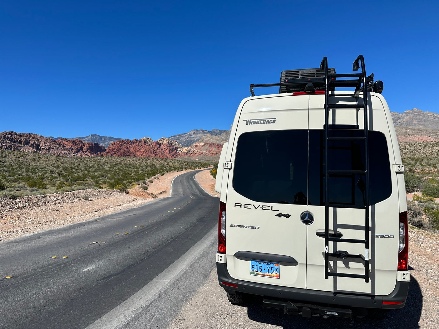 A Winnebago Revel 4x4 rv rental parked on a winding desert road, surrounded by stunning red rock mountains under a clear sky.