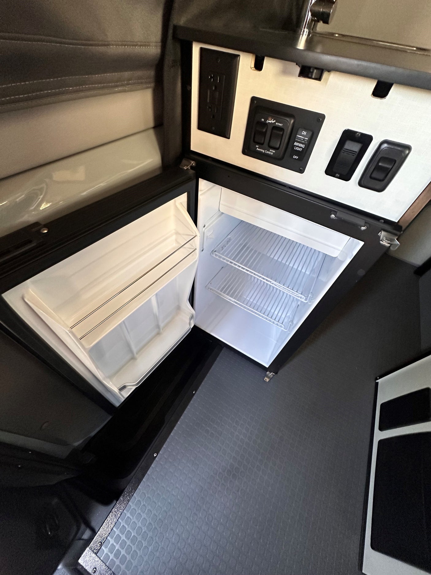 Compact RV rental interior: open mini-fridge, control panel, electrical outlet, and textured floor visible.
