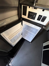 Compact RV rental interior: open mini-fridge, control panel, electrical outlet, and textured floor visible.