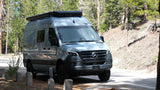This light gray Mercedes-Benz Revel camper van, an ideal RV rental, is parked for an off-road adventure in a sunny pine forest.
