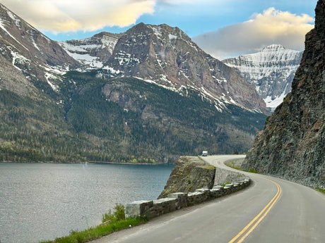 An RV travels a winding lakeside road, framed by snow-capped mountains and evergreen forests. Ideal for an RV rental trip.