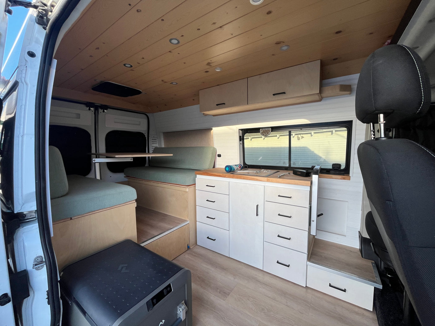 Step into this bright RV rental camper van, featuring a wood ceiling, sage green cushioned seating, kitchen, and smart storage solutions.