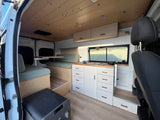 Step into this bright RV rental camper van, featuring a wood ceiling, sage green cushioned seating, kitchen, and smart storage solutions.